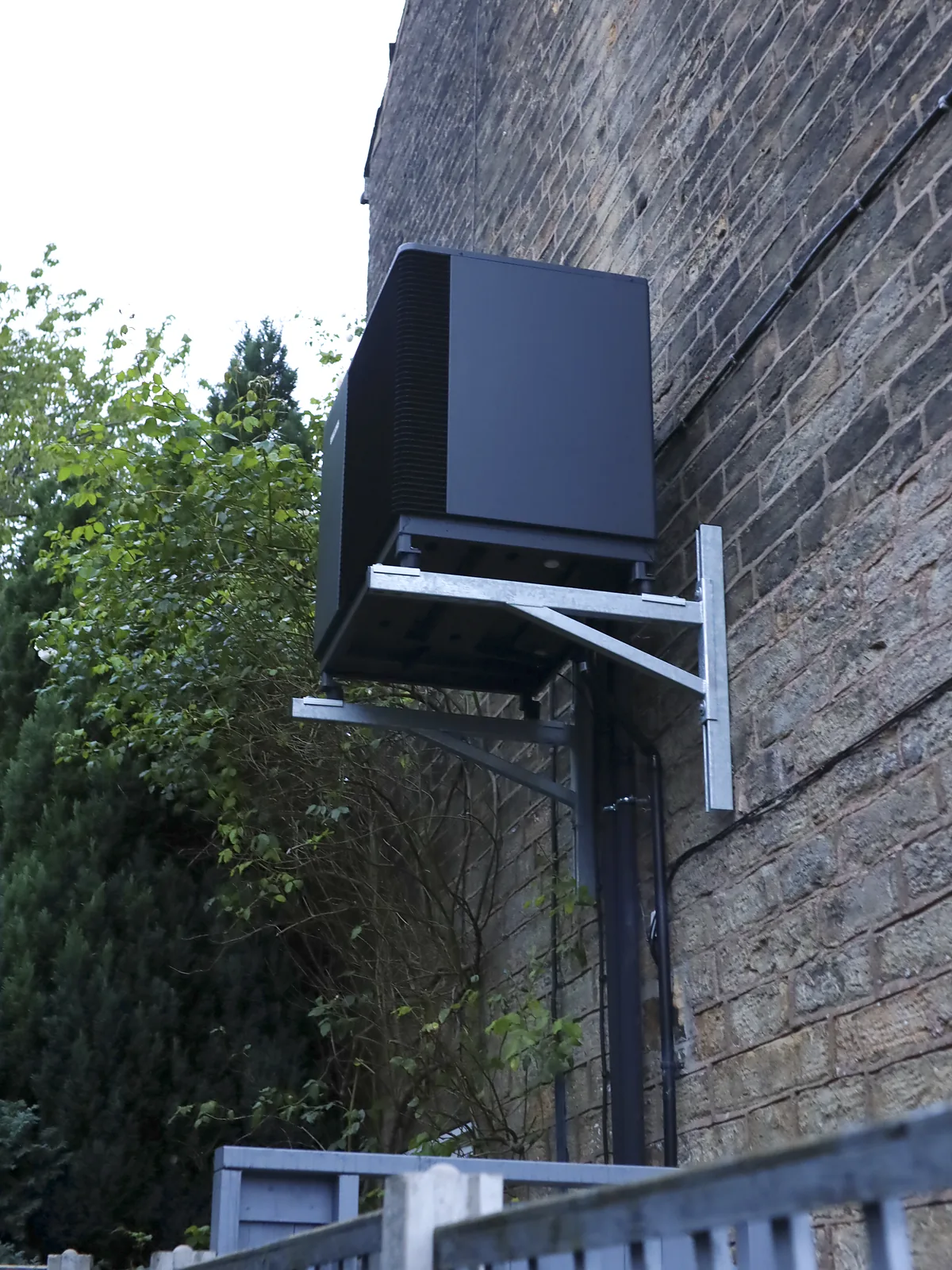Air source heat pump on a galvanised wall bracket high on a stone wall, seen from the garden below with trees and a timber fence