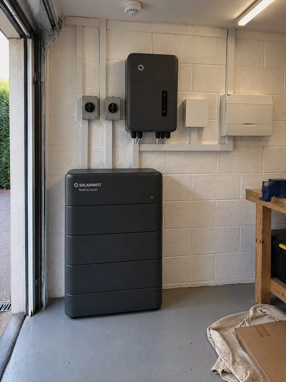 Illustration of a SolarWatt home battery in the double garage of a detached house, the inverter, isolators and consumer unit beside it in white trunking