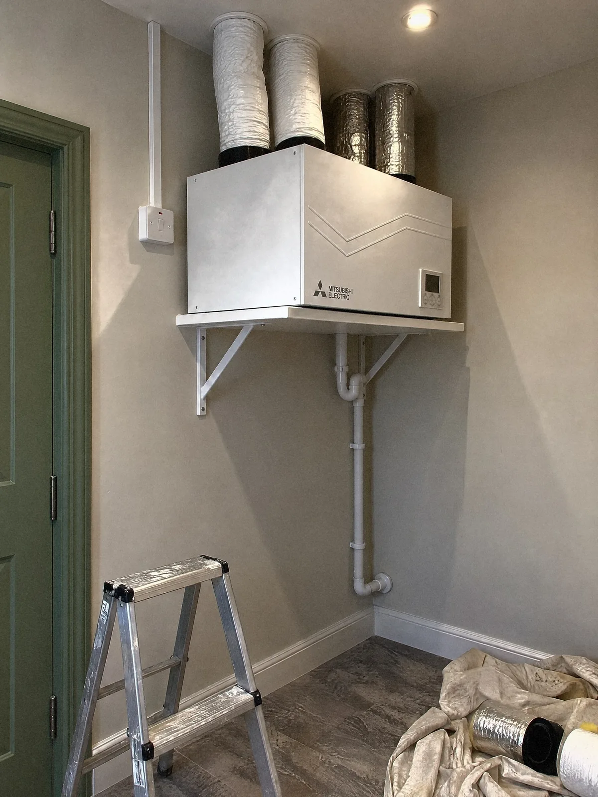 Illustration of a white Mitsubishi Electric heat recovery unit on a bracketed shelf in a utility room, its four insulated ducts rising into the ceiling