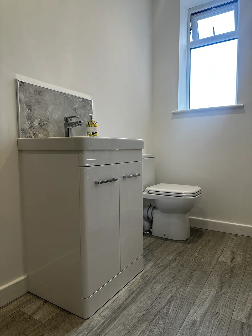 A finished cloakroom with a white vanity unit, a grey marble-effect splashback and a close-coupled toilet under a window.