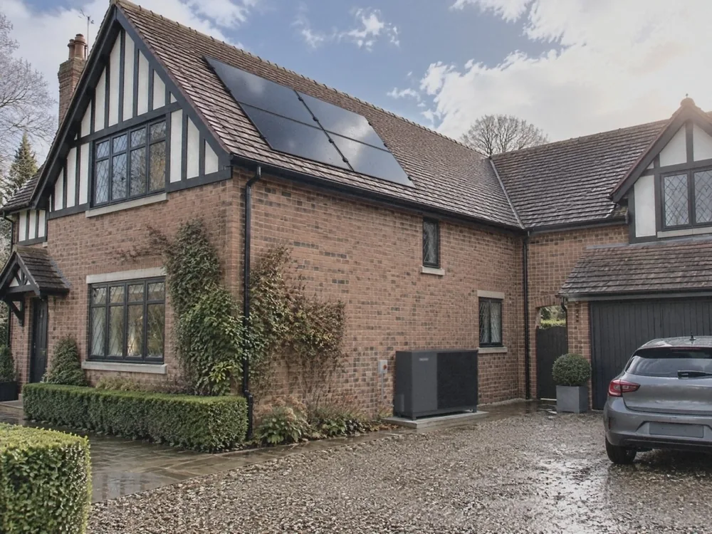 Illustration of a red-brick and half-timbered detached house just after rain, four black solar panels on its clay-tiled roof and a dark grey heat pump on a slab by the wet gravel drive