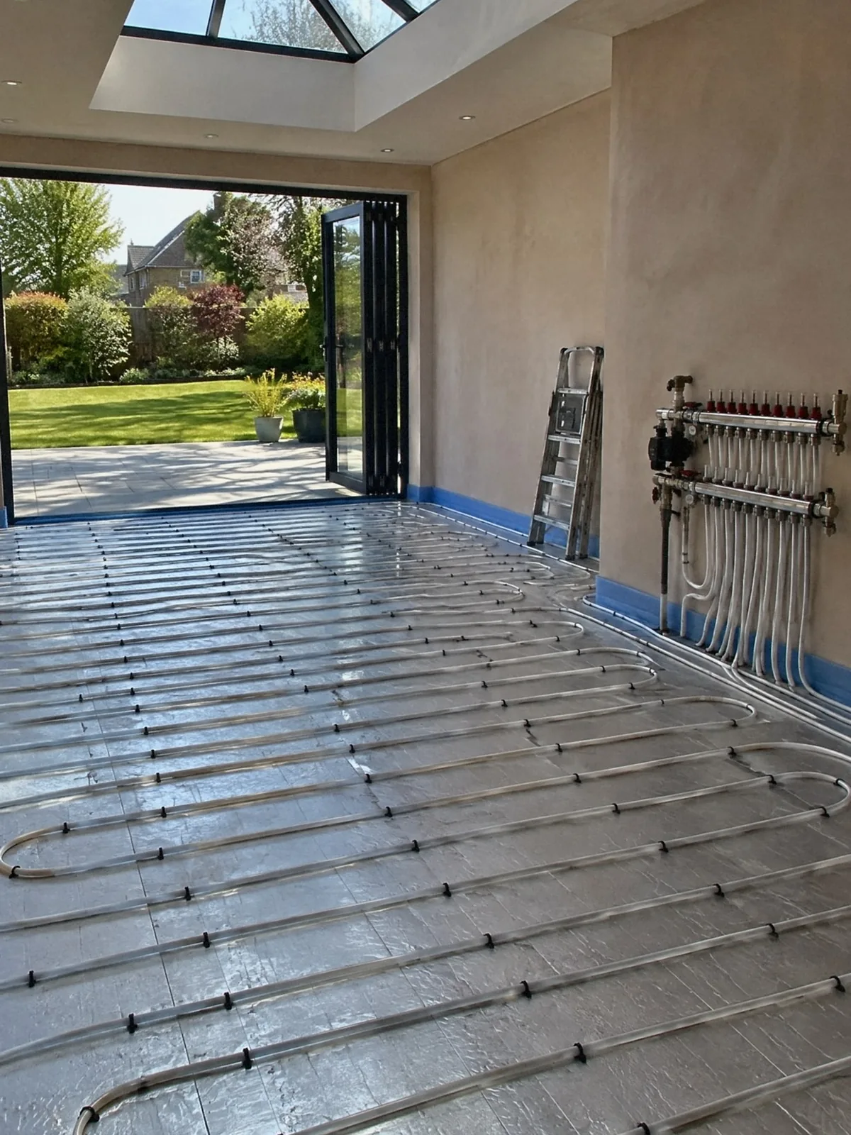 Illustration of underfloor heating pipes clipped in loops across the floor of a new extension, the manifold on the wall and a garden through open bifold doors