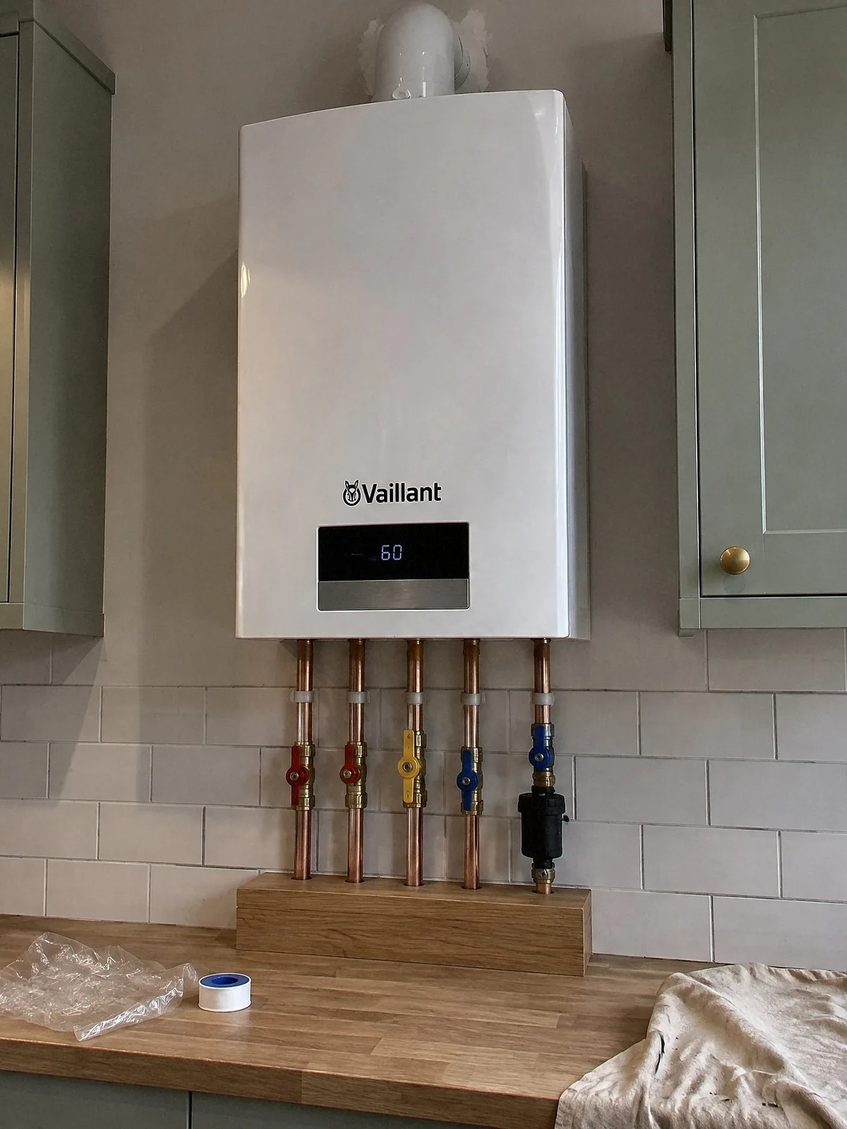 Illustration of a new white Vaillant combi boiler between sage shaker wall cupboards in a kitchen, its copper pipework running into an oak pipe cover on the worktop