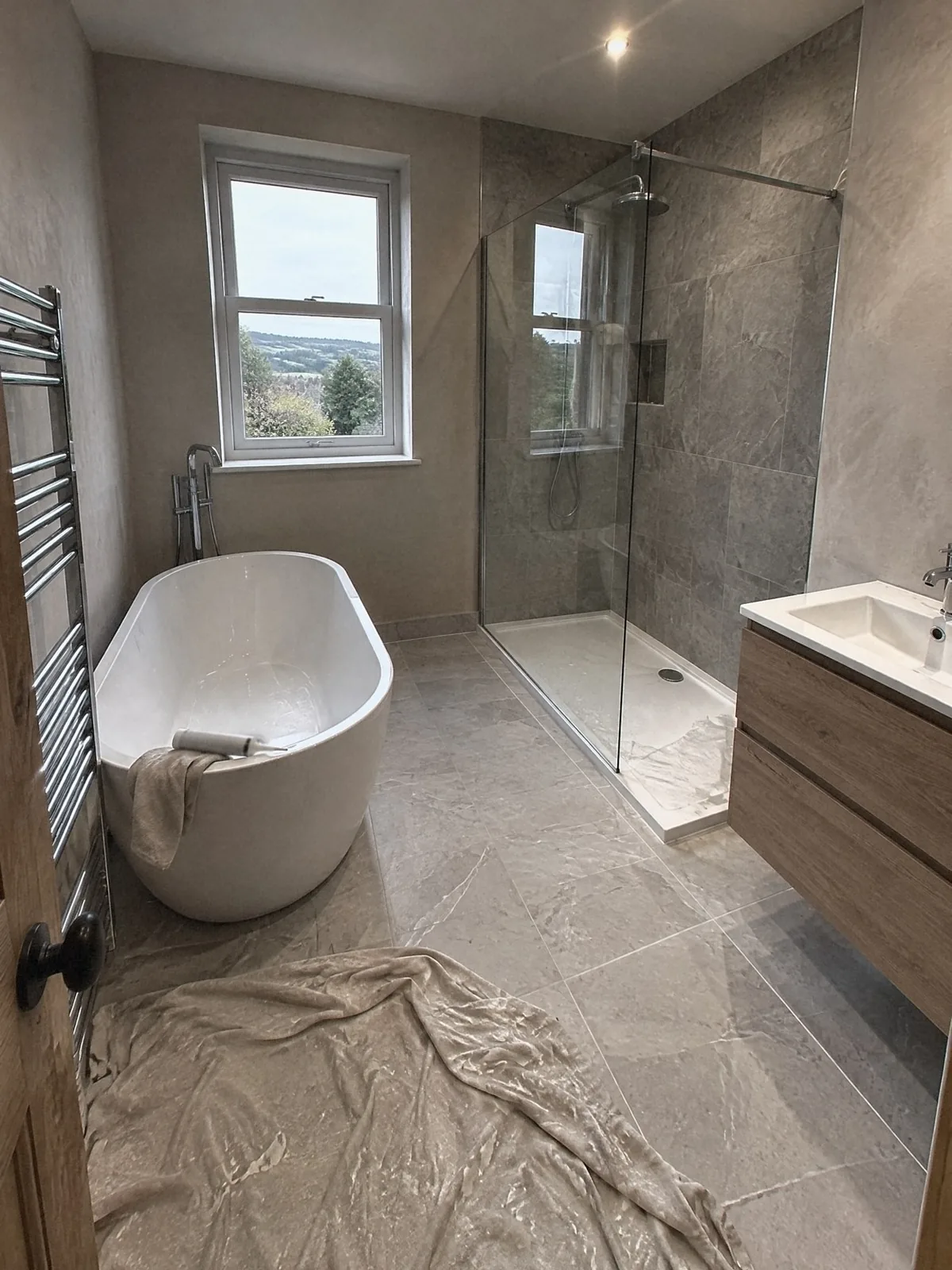 Illustration of a just-finished bathroom with a freestanding bath, a walk-in shower and an oak vanity, a dust sheet still on the floor and green hills through the window