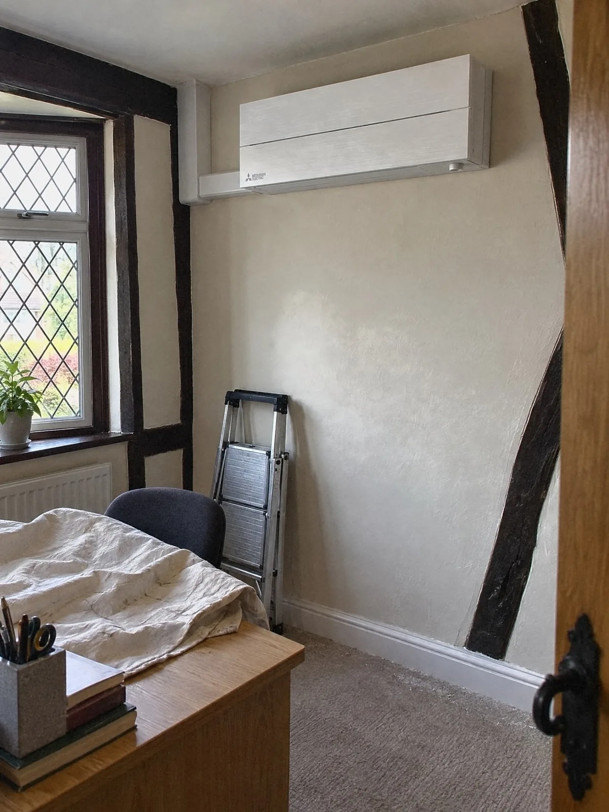Illustration of a new white Mitsubishi Electric air conditioning unit high on the wall of a home office with leaded windows, a dust sheet over the desk and a stepladder by the wall