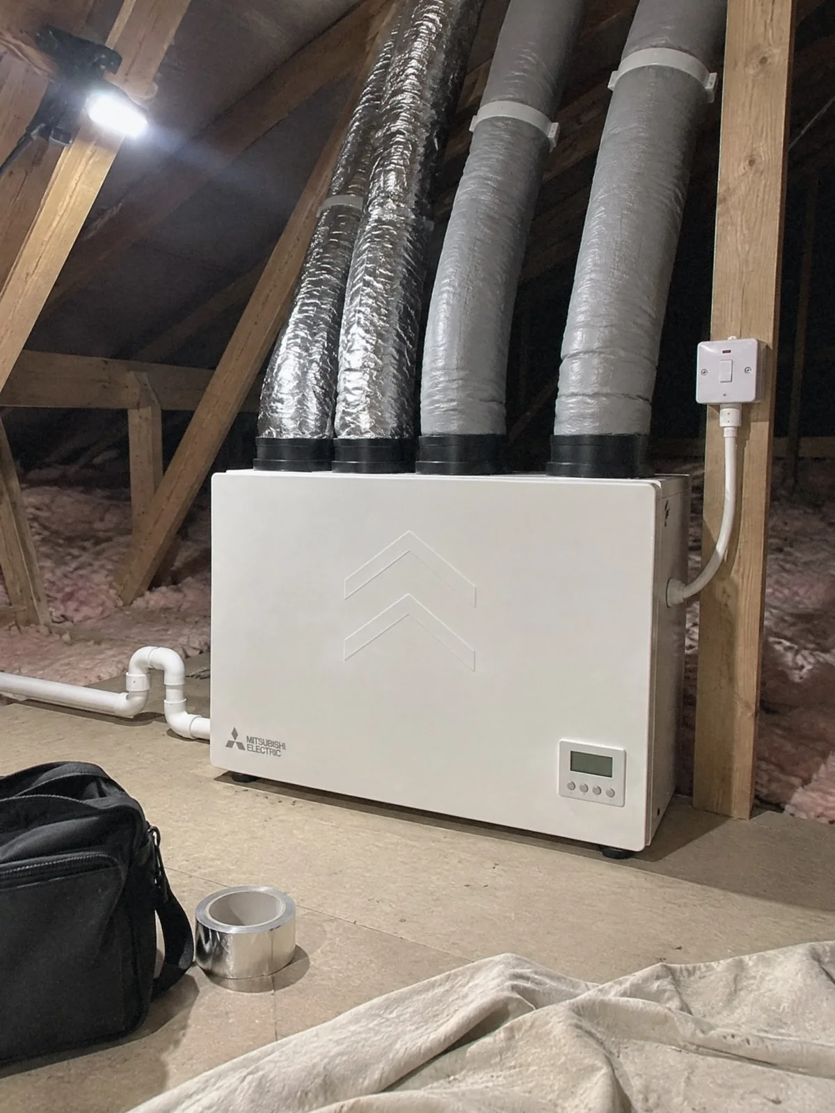 Illustration of a white Mitsubishi Electric heat recovery unit on the boarded floor of a loft, foil-wrapped and grey insulated ducts rising from its top and a fused spur on the timber beside it