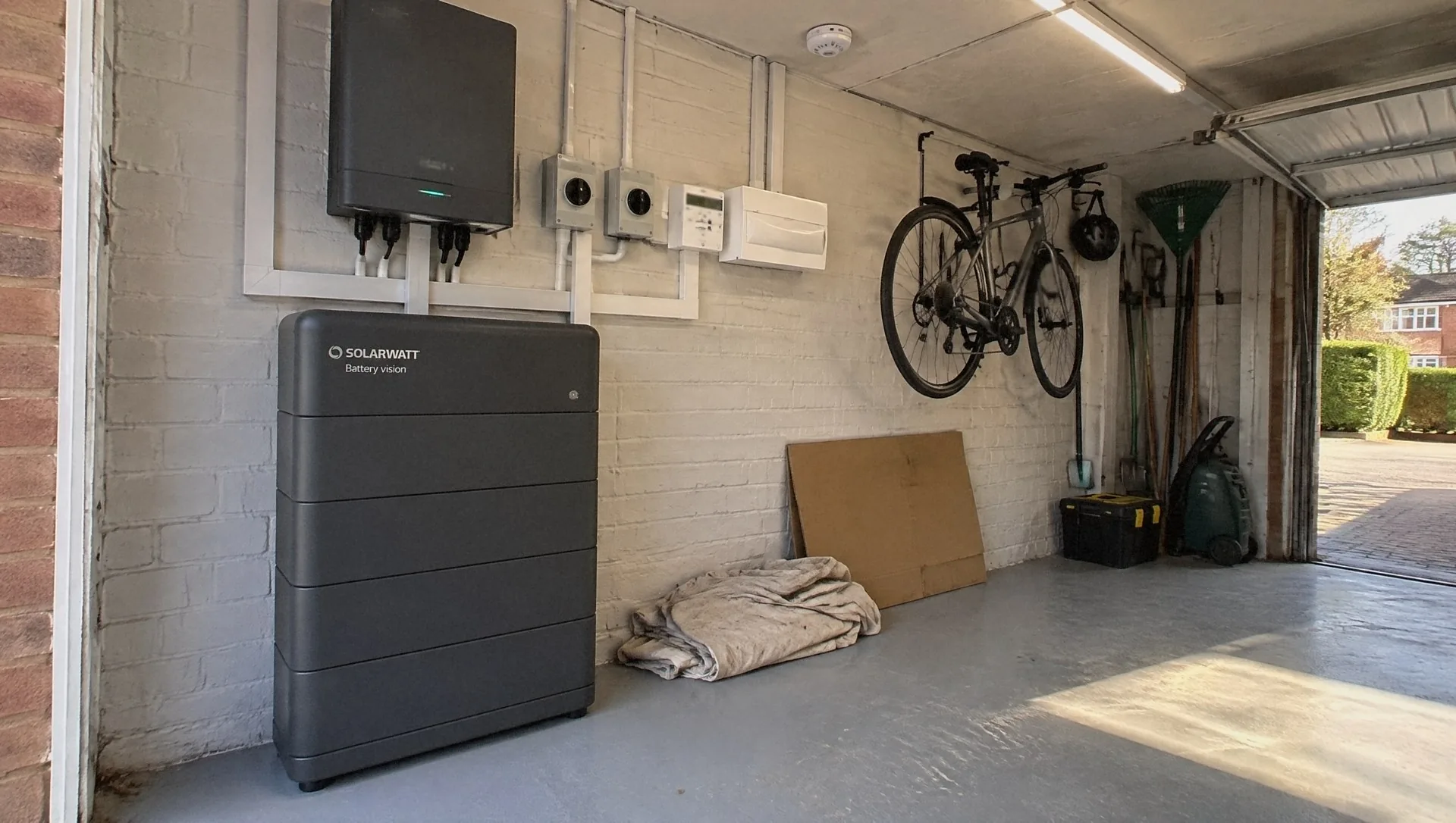 Illustration of a SolarWatt home battery in a detached garage, its inverter, isolators, meter and consumer unit above it in white trunking, a bike on the wall and sunlight through the open door