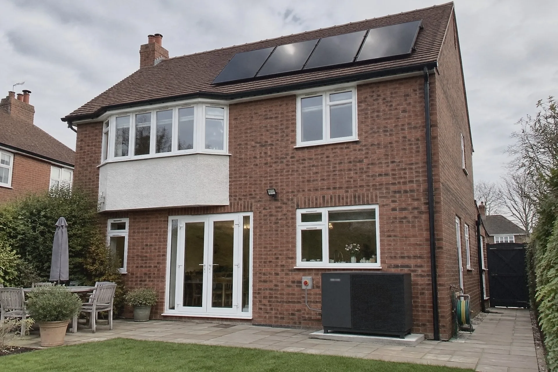 Illustration of the back of a red-brick detached house on a cloudy day, four black solar panels on its tiled roof and a dark grey heat pump on a slab by the patio