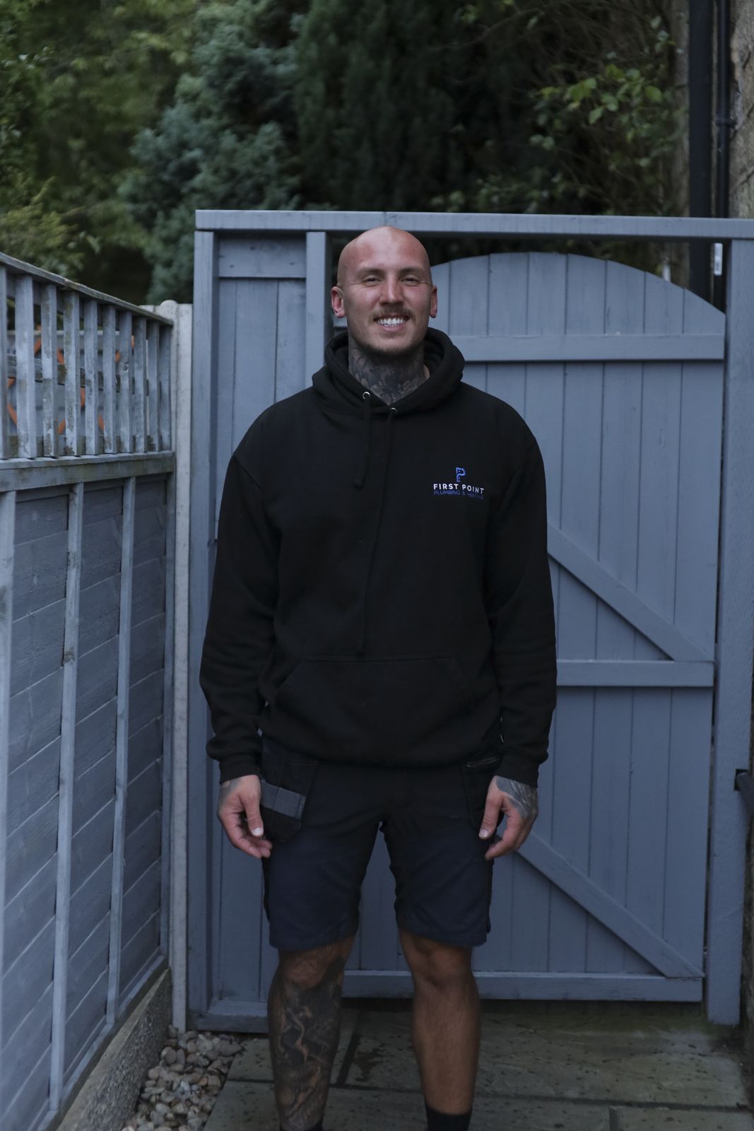 First Point engineer in a black branded hoodie standing in the open garden gate of a customer's property, grey larch fence panels and a conifer behind him