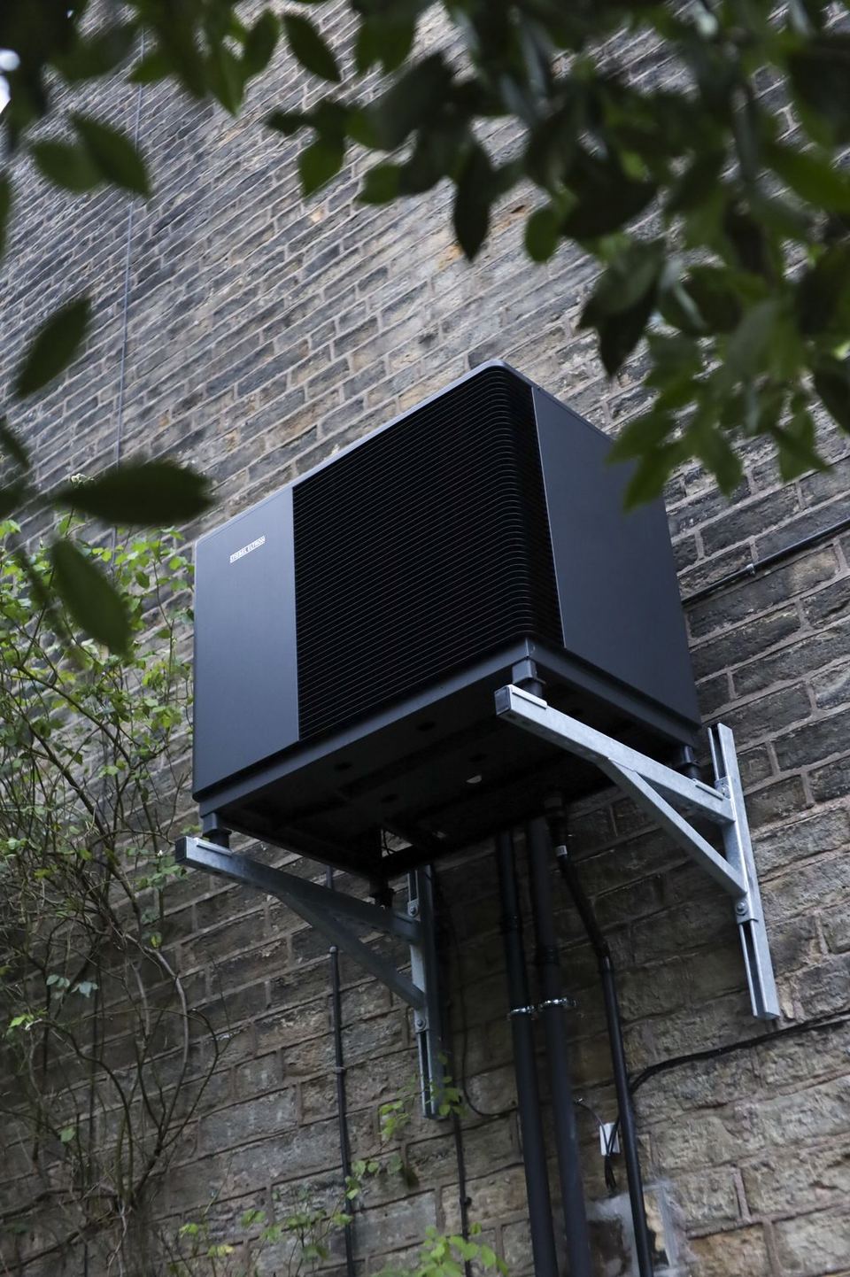 Air source heat pump on a wall bracket, seen from below against a stone wall with leaves framing the shot