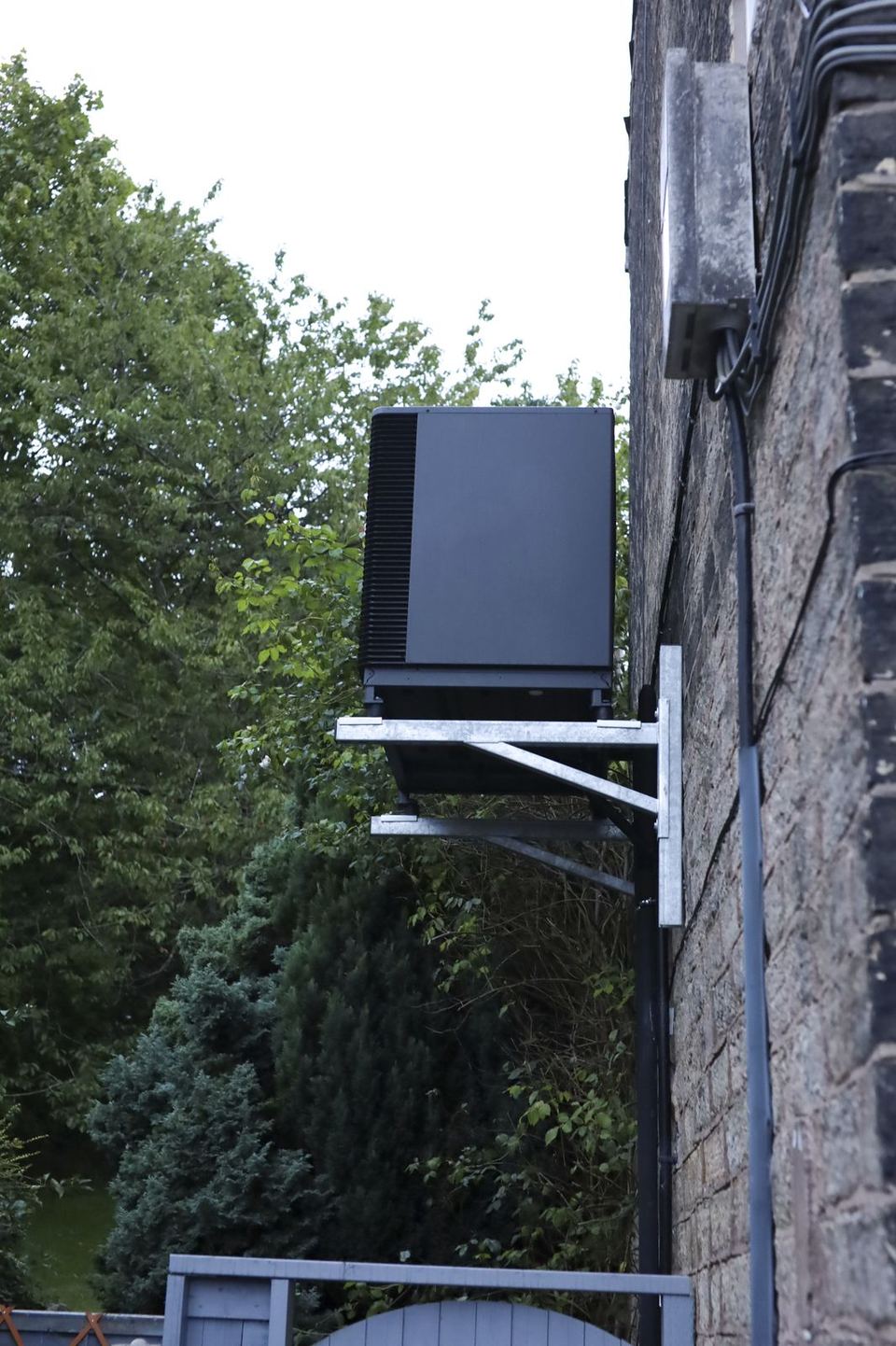Air source heat pump on its wall bracket seen from further back, garden trees to one side and a timber gate below it