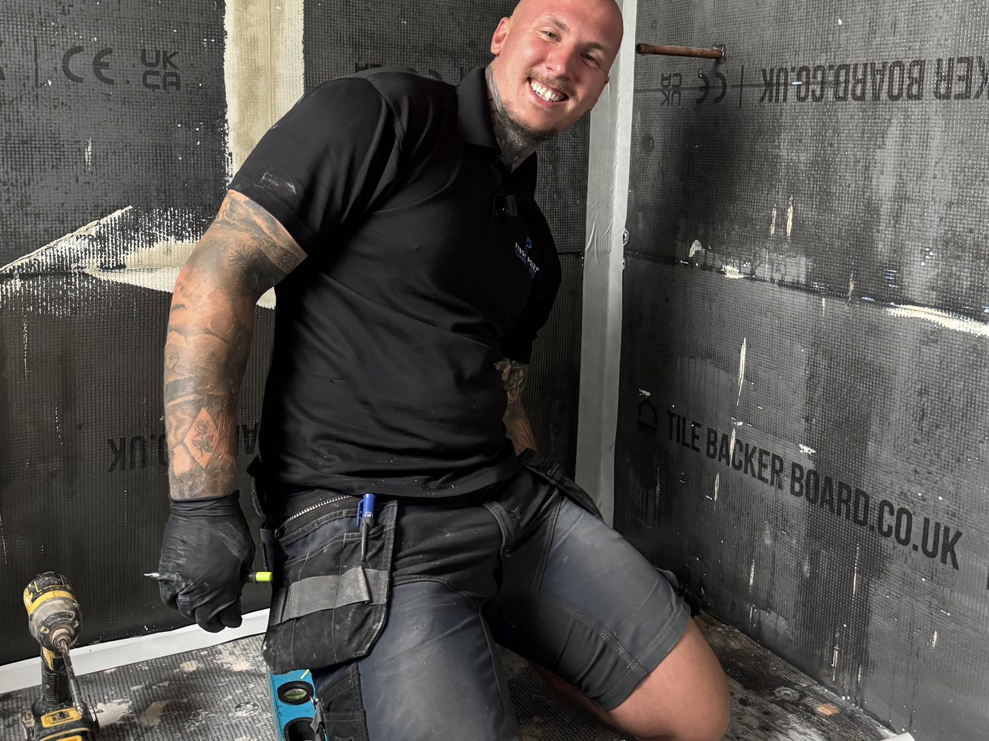 First Point engineer kneeling in a stripped shower enclosure lined with tile backer board, a cordless drill and a spirit level on the floor beside him