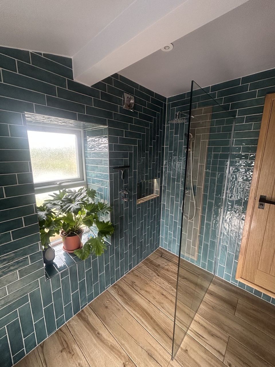Wet room in teal green metro tile, a glass screen, chrome rain head and a potted plant on the window sill