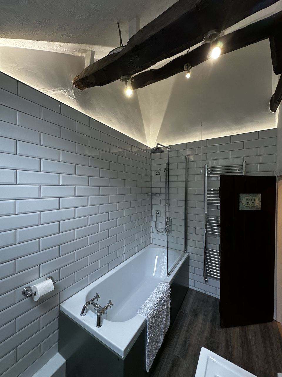 Period cottage bathroom in white metro tile under a dark timber beam, a bath with a green panel and a chrome rain head
