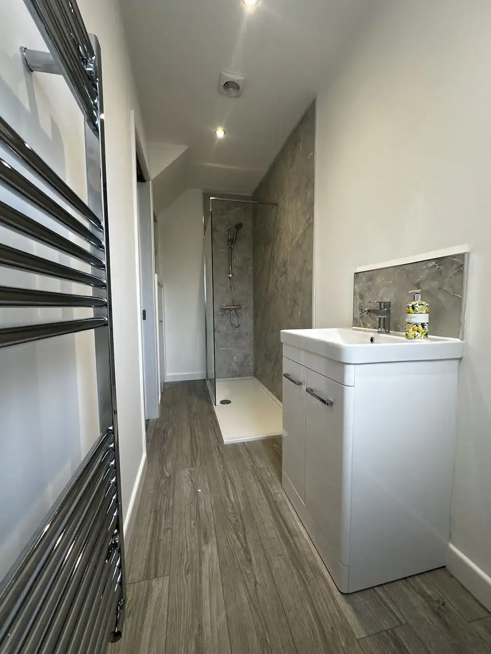 Narrow shower room with a chrome ladder towel radiator in front, grey wood-effect flooring, a white vanity and a grey marble-effect walk-in shower