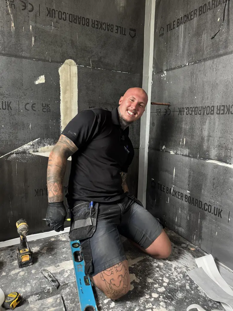 First Point engineer kneeling in a stripped shower enclosure lined with tile backer board, a cordless drill and a spirit level on the floor beside him