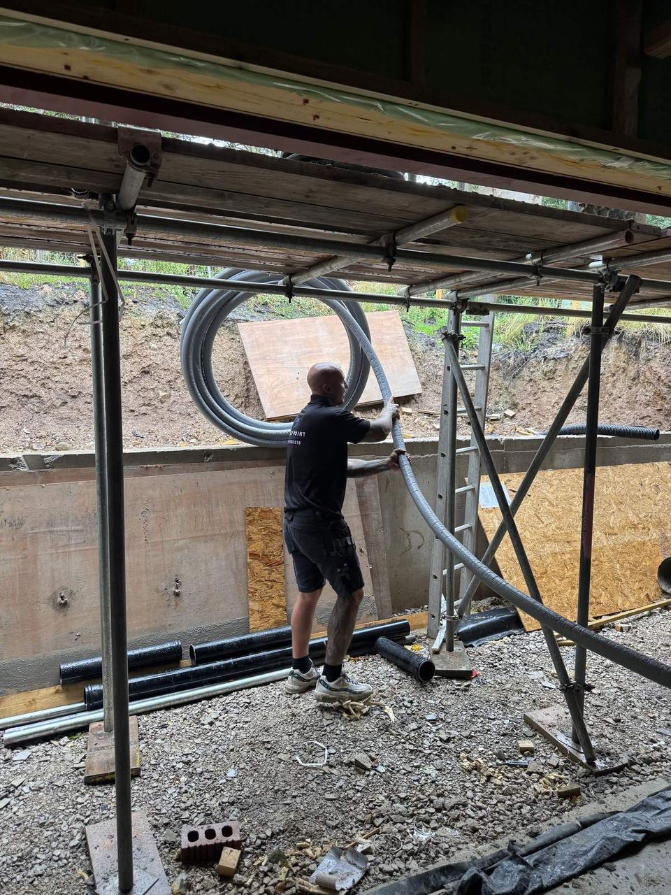 Engineer feeding grey flexible ducting over a scaffold lift into a part-built house’s timber floor structure