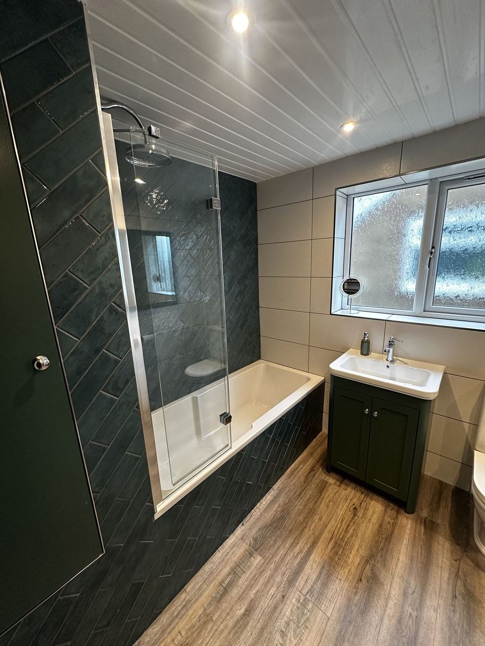 Bathroom in dark green herringbone tile, a bath with a chrome rain head behind a glass screen and a green vanity