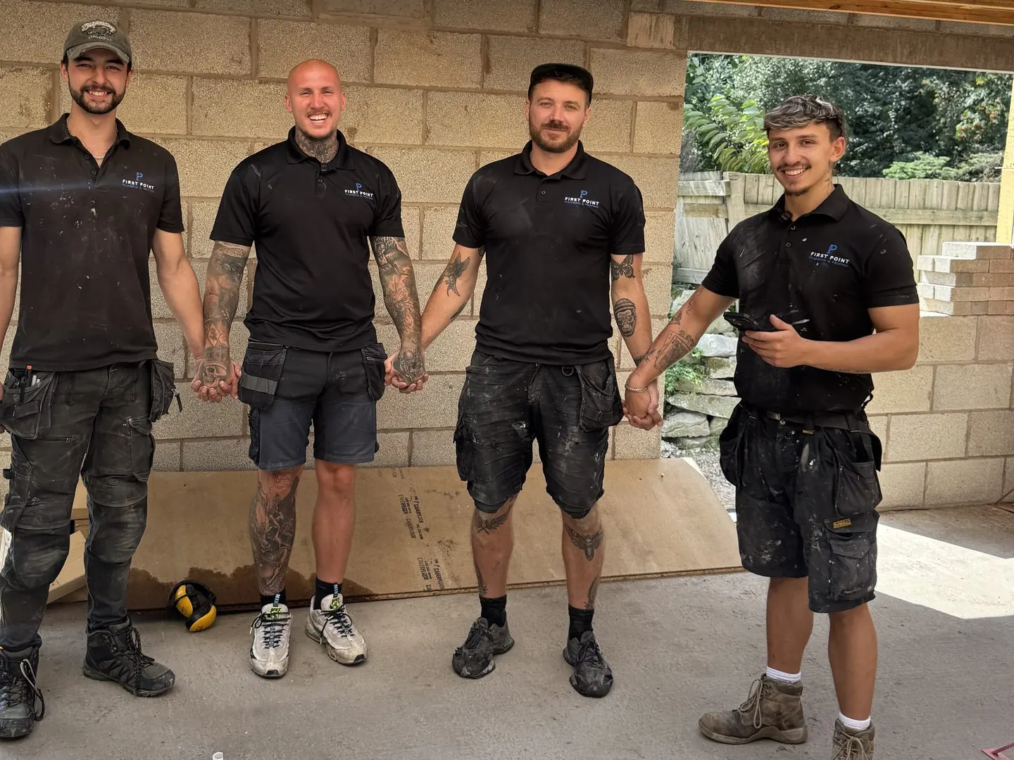 Four First Point engineers in black polo shirts holding hands and laughing under a new timber roof, fresh blockwork behind them