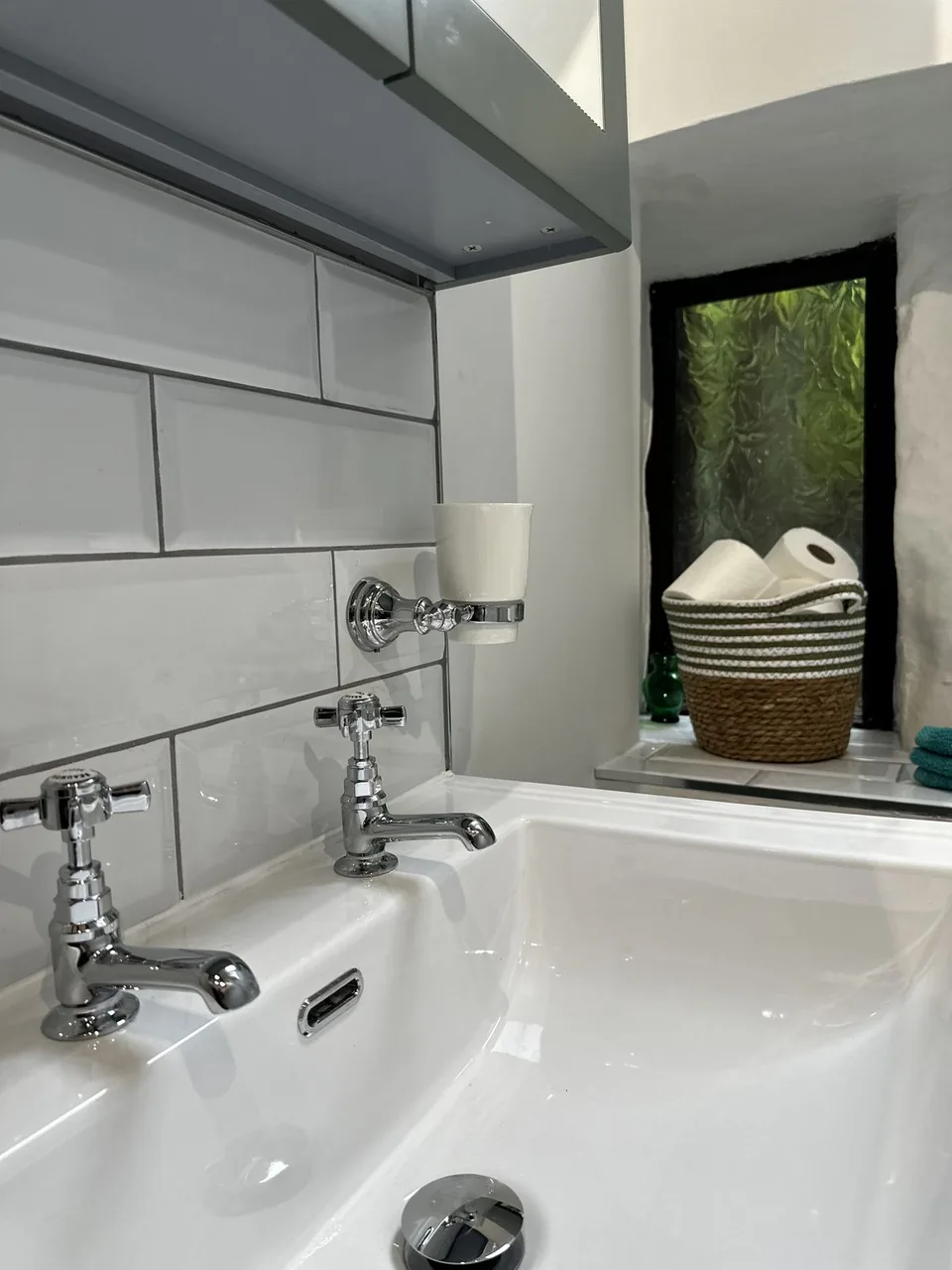 Two chrome crosshead pillar taps on a white basin against white bevelled metro tile, with a deep whitewashed window reveal and greenery outside