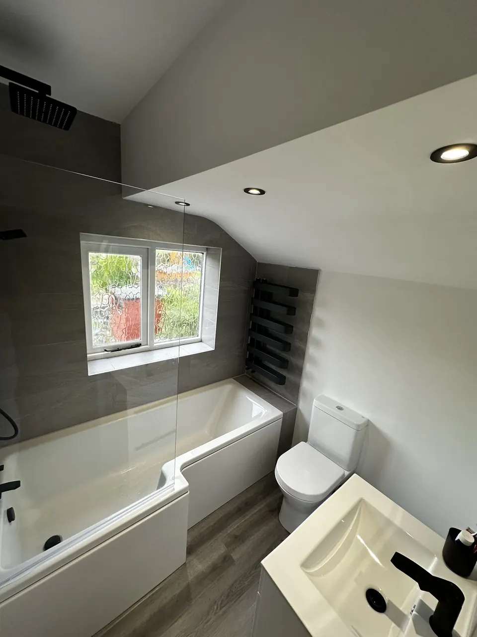 Small bathroom under a sloping ceiling with an L-shaped shower bath, grey stone-effect tiling, a black towel radiator and a white basin with a black tap