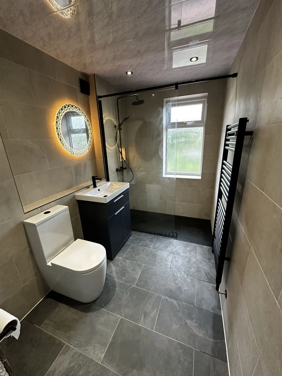 Shower room with a backlit round mirror, navy vanity, matt black framed walk-in screen and black ladder towel radiator