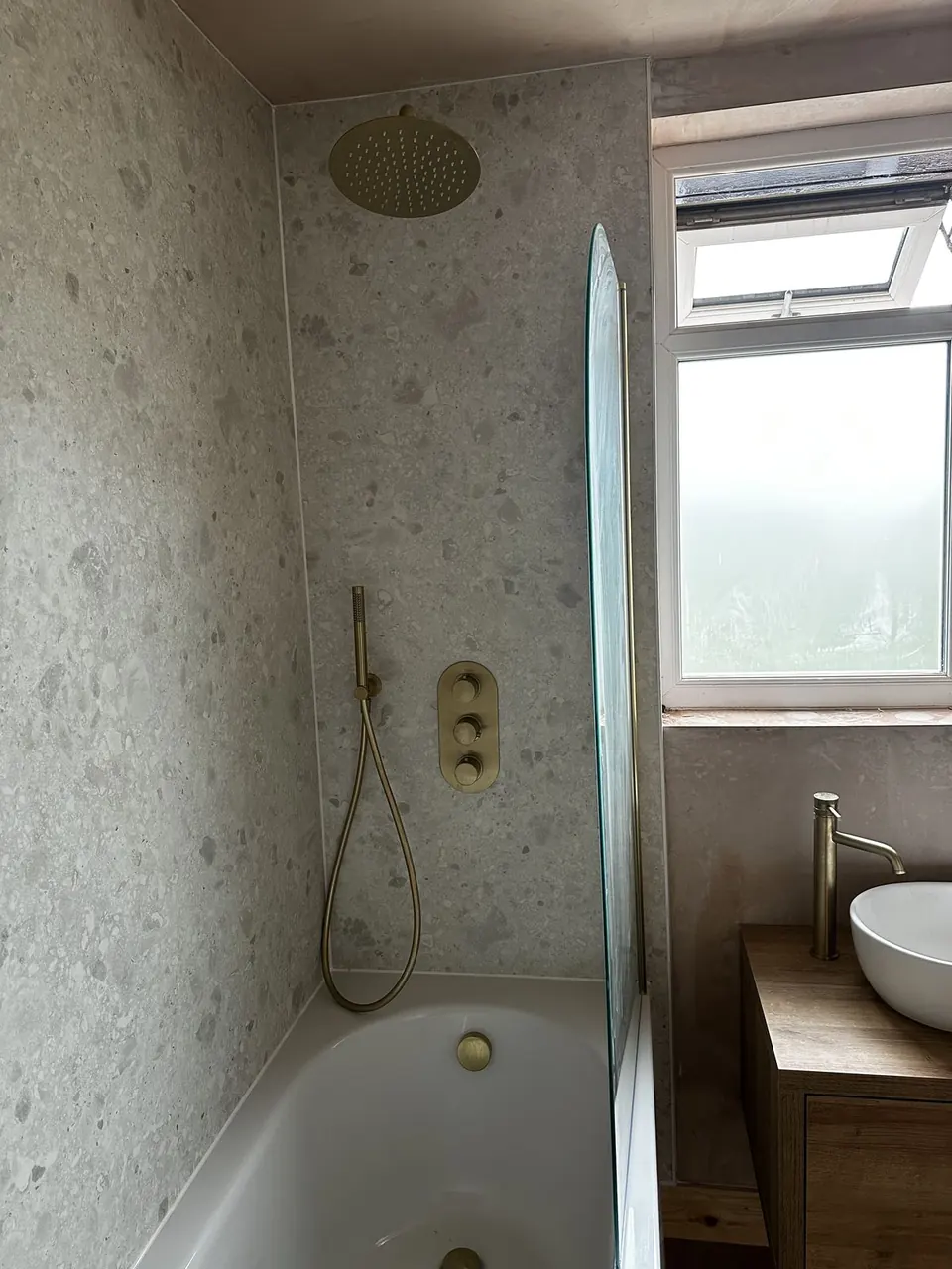 Bath with terrazzo-effect wall panels, a brushed brass rain head and valve, and an oak-topped vanity with a vessel basin, one wall still bare plaster