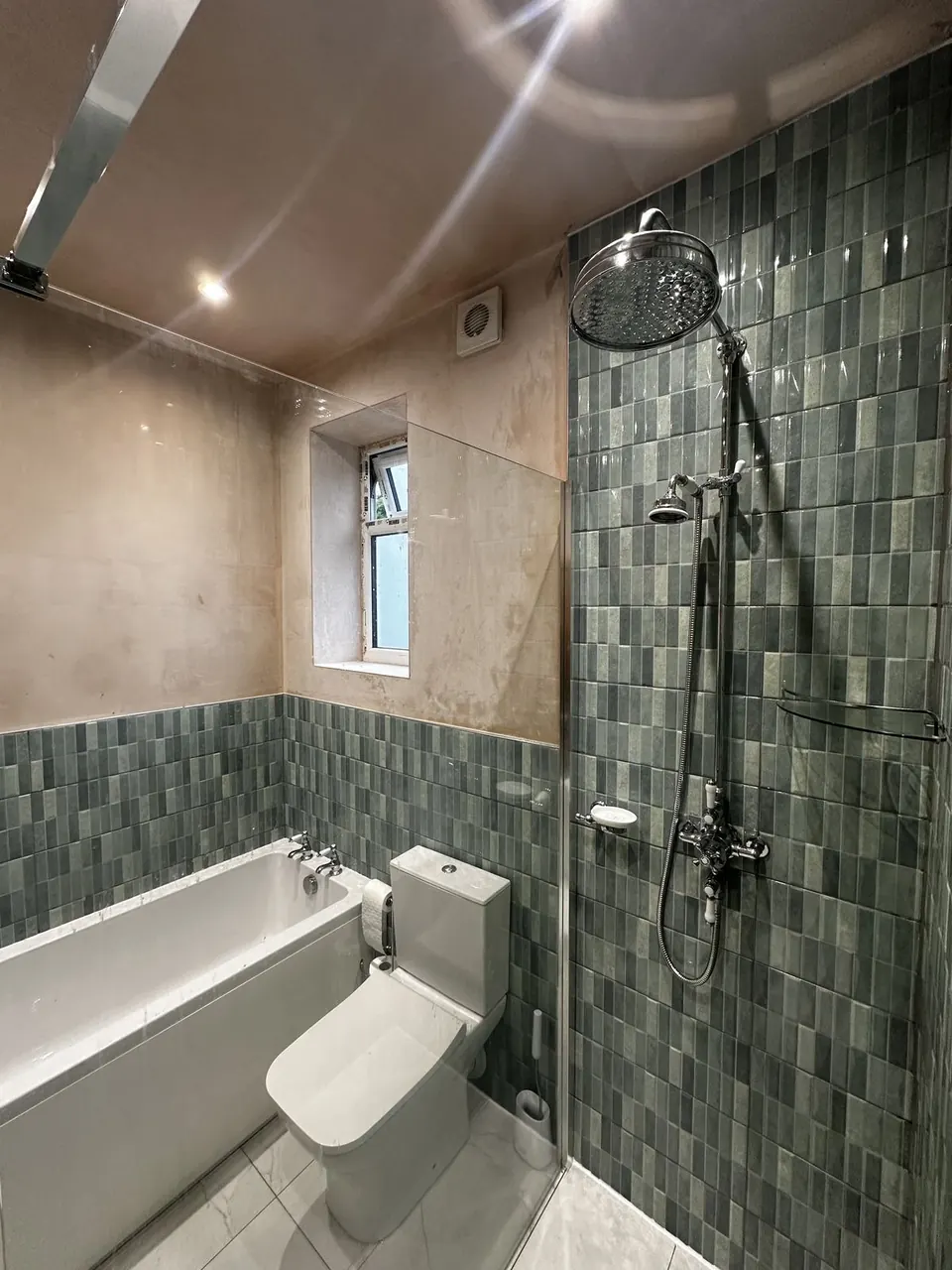 Bath with a chrome exposed shower valve and rain head against green-grey mosaic strip tile, the upper walls still bare plaster