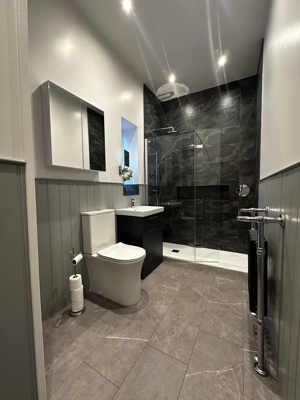 Shower room with pale grey panelling, a black vanity, a period-style radiator and a walk-in shower lined in dark grey marble-effect tile